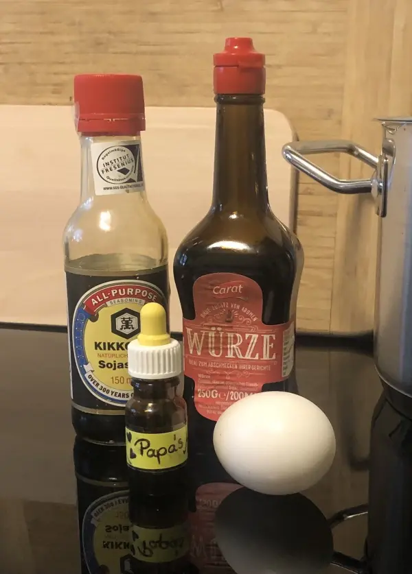 Eine Selection von Essenzen, einschließlich Sojasauce und Würze, steht neben einem Ei auf einer schwarzen Oberfläche.