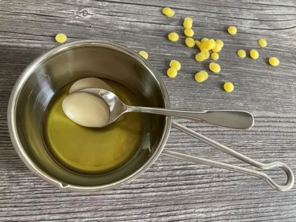 Ein silberner Topf mit Öl und einer Löffel wartet auf die Zubereitung von Lippenbalsam mit Honig, umhüllt von Wachskügelchen.