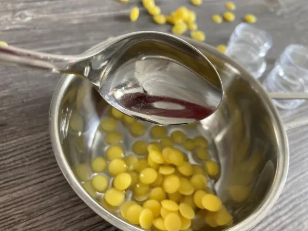 Ein Metalllöffel hält etwas über einem Topf mit geschmolzenen Wachsperlen, während im Hintergrund leere Behälter stehen.