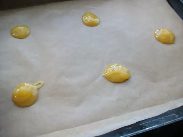 Auf einem Backblech liegen kleine Tropfen einer gelben Masse auf Backpapier, bereit zum Backen von Gebäck.