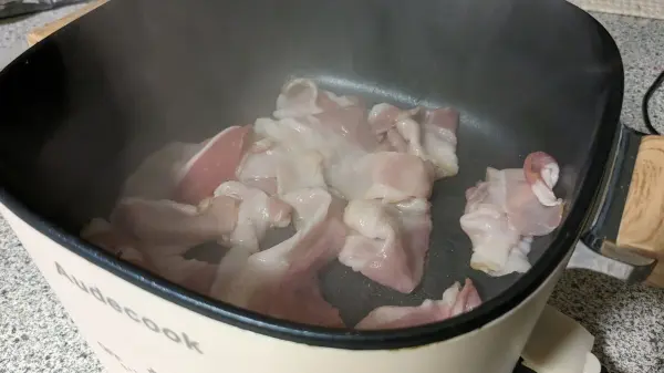 Ein Stück Speck brät in einer Heißluftfritteuse, während Dampf aufsteigt und die Umgebung leicht vernebelnd wirkt.