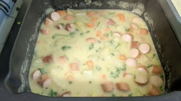 Ein cremiger Gemüseeintopf mit Würstchen wird in einer Heißluftfritteuse zubereitet, umgeben von einer modernen Küche.