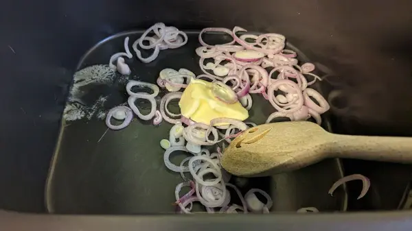 In der Heißluftfritteuse schwitzen geschnittene Zwiebeln und Butter, während ein Holzlöffel zum Rühren bereitliegt.