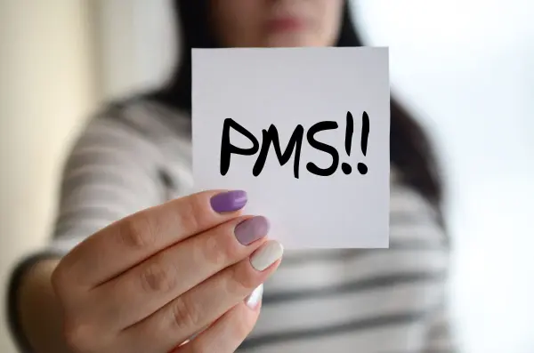 Eine Hand hält einen Zettel mit der Aufschrift „PMS!!“ vor einem verschwommenen Hintergrund, was die Thematik des PMS verdeutlicht.