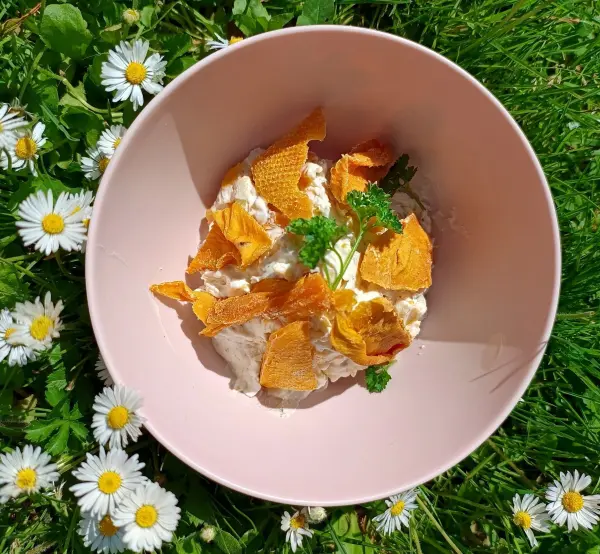 In einer rosa Schüssel liegt Frischkäse, garniert mit Mangostreifen und frischer Petersilie, umgeben von Gänseblümchen.