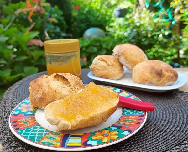 Ein knuspriges Brötchen mit Ananas-Zitronen-Gelee liegt auf einem bunten Teller, während weitere Brötchen im Hintergrund stehen.