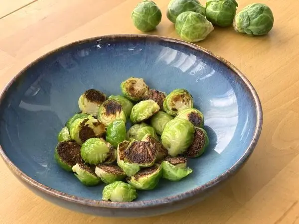 Rosenkohl, goldbraun gebraten, liegt in einer blauen Schüssel auf einem Holztisch. Im Hintergrund liegen rohe Rosenkohlköpfe.