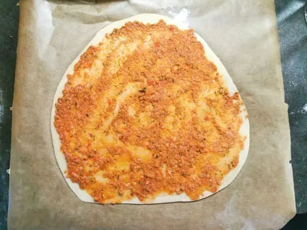 Auf einem Backpapier liegt ein roher Lahmacun in runder Form, belegt mit einer würzigen Mischung aus Paprika, Tomatenmasse und Hackfleisch .