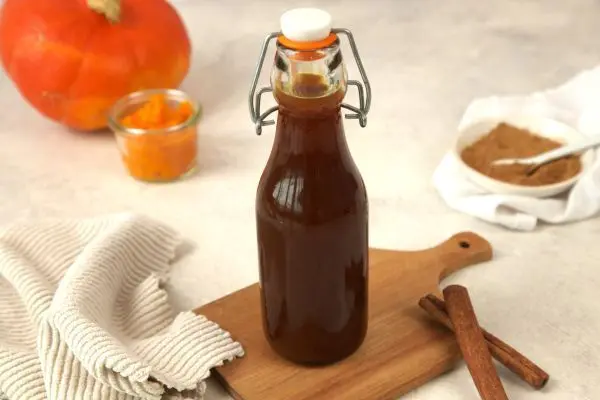 Braune Flasche mit Pumpkin Spice Sirup steht auf einem Holzbrett, umgeben von Kürbis, Gewürzen und Küchentuch.