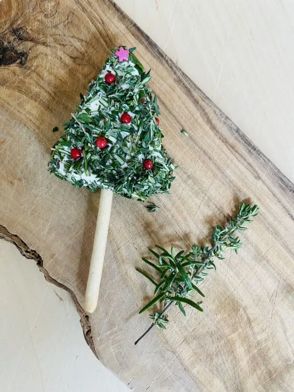 Ein Schmelzkäse-Tannenbaum, verziert mit Kräutern und rotem Pfeffer, liegt auf einem rustikalen Holzbrett mit Thymianzweig.