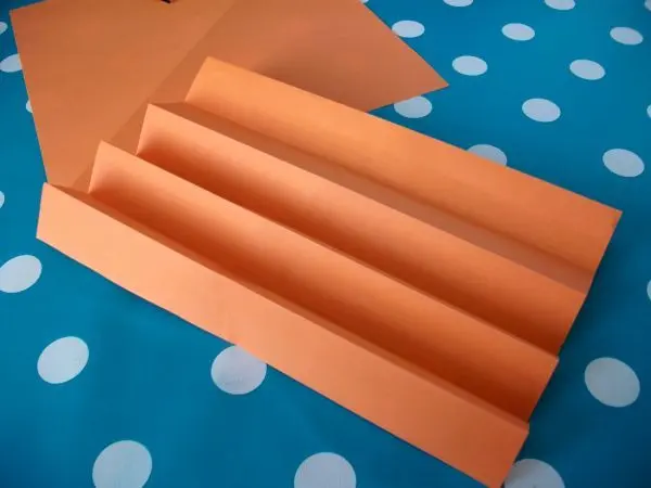 Orangefarbenes Papier liegt wie eine Ziehharmonika gefaltet auf einem blauen, gepunkteten Tischtuch und bildet die Grundlage für Papiersterne.