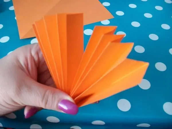 Eine Hand hält ein gefaltetes orangefarbenes Papier auf einem blauen Tisch mit weißen Punkten.