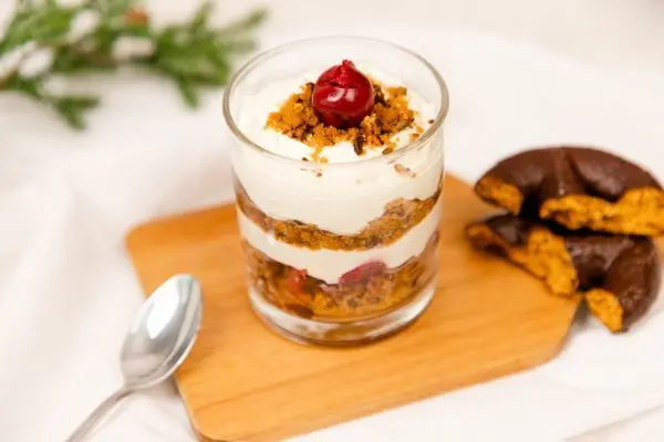 Ein Tiramisu mit Lebkuchen im Glas, geschichtet mit Creme und Kirschen auf Holzbrett; daneben liegen halbierte Lebkuchen.