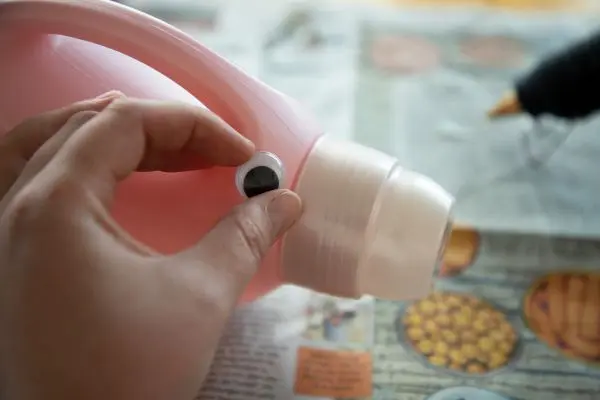 Eine Hand klebt ein Wackelauge auf eine rosa Waschmittelflasche, die auf einer Zeitung liegt.