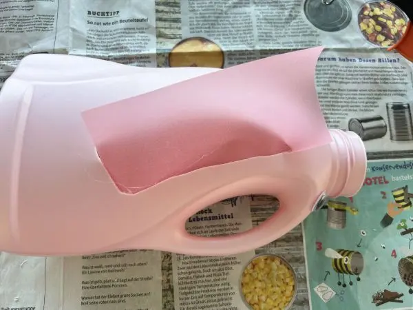 Eine alte Waschmittelflasche wird auf Zeitungspapier liegend für ein Laternenbastelprojekt mit rosa Transparentpapier beklebt.