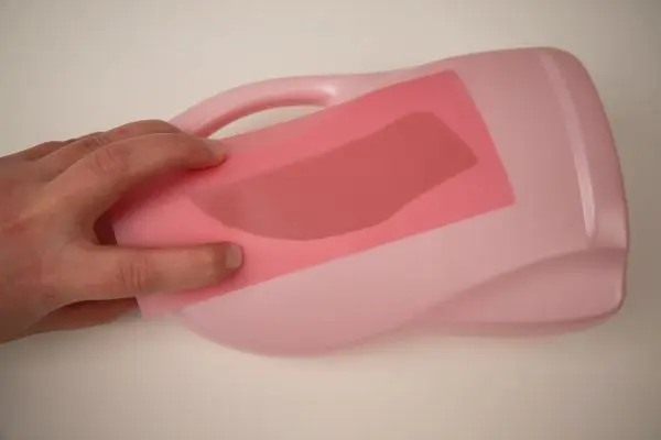 Eine Hand hält rosa Transparentpapier auf eine leere, rosa Waschmittelflasche. Die Flasche liegt auf einem weißen Untergrund.