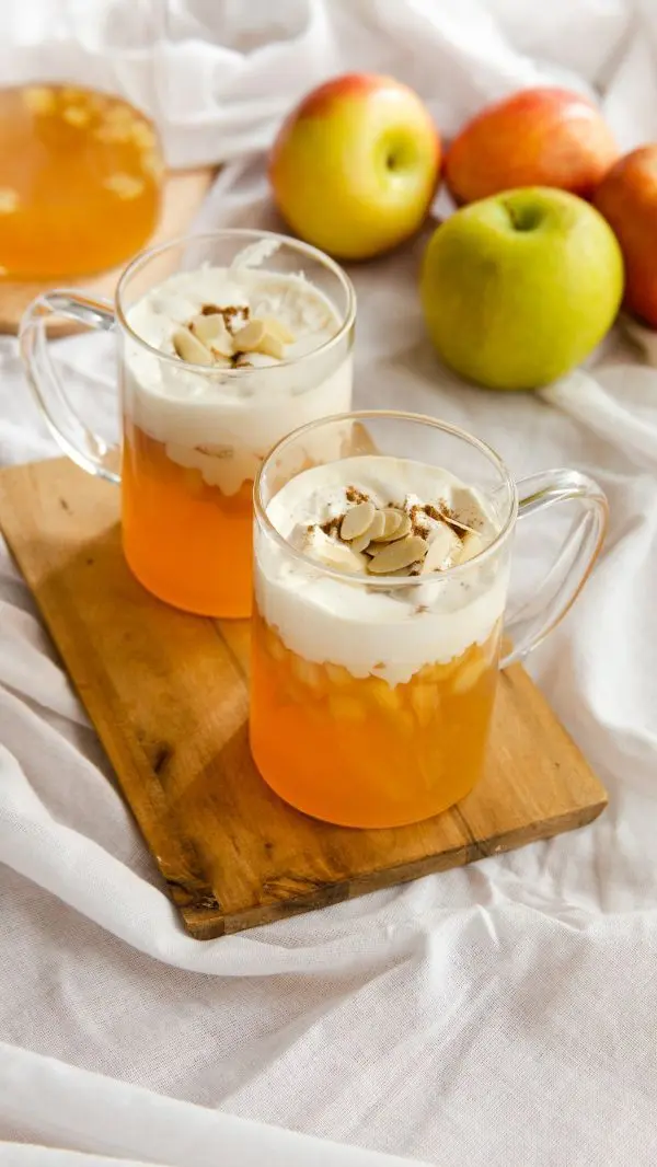 Zwei Gläser Apfelpunsch (ohne Alkohol) mit Vanille-Sahne stehen auf einem Holzbrett; dahinter liegen frische Äpfel.