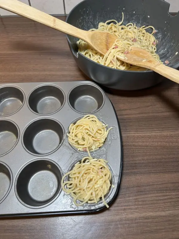 Verteile die Spaghettimischung jetzt auf die zwölf Muffinförmchen. Nimm dabei nur wenige Nudeln zwischen das Salatbesteck, sonst werden die Förmchen zu voll. Außerdem kleckerst du so auch weniger.