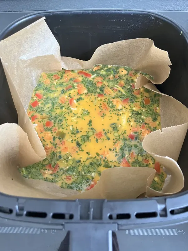 Omelette aus dem Airfryer vor dem Garen &ndash; fl&uuml;ssige Eiermasse mit Schnittlauch, Fr&uuml;hlingszwiebeln und Paprika im Airfryer-Korb auf Backpapier.