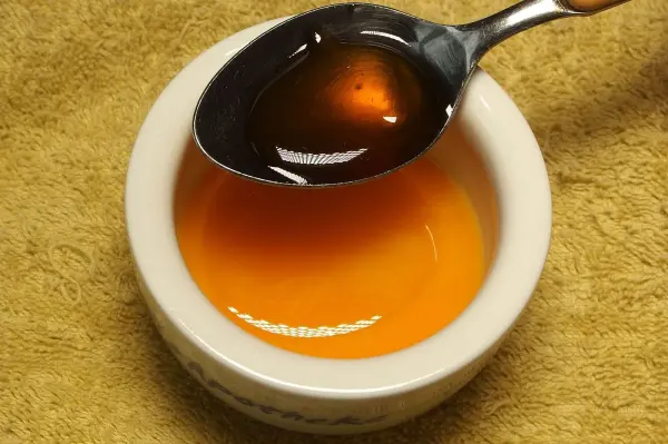 Ein Löffel Honig wird über einer kleinen Schale mit Honig gehalten, auf einem weichen, gelben Untergrund.