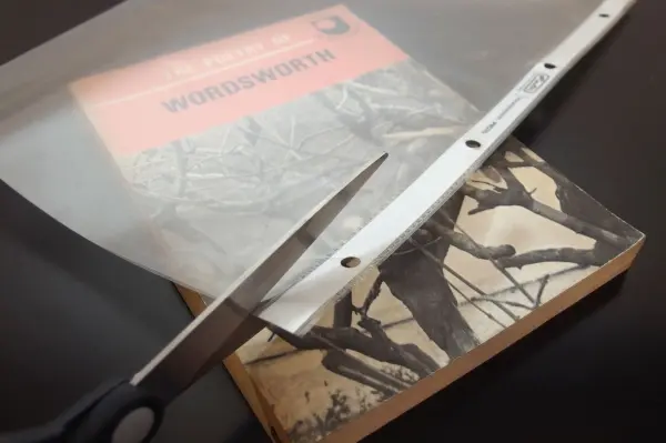 Eine Schere schneidet durch eine transparente Bucheinbandfolie, die auf einem Buch liegt. Der Hintergrund ist dunkel.