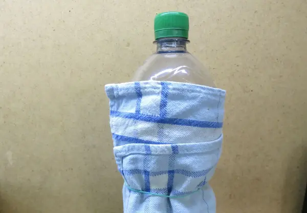 Eine Plastikflasche ist eng in ein blau-weiß kariertes Küchentuch gewickelt, steht vor einem neutralen Hintergrund.