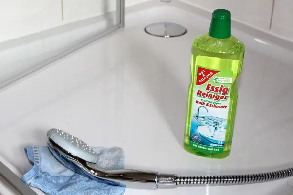In einer Dusche steht eine Flasche Essigreiniger neben einem Reinigungstuch und einer Duschbrause auf dem fliesenboden.