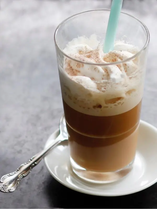Ein Glas Frappe mit Schaumkrone steht auf einem Unterteller, ein Löffel daneben, in einem neutralen Umfeld.