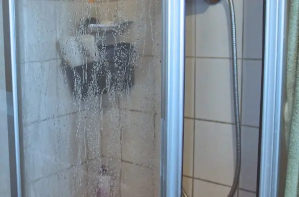 Eine Glas-Duschkabine wird mit Wasser besprüht und hat Schaumrückstände, steht in einem gefliesten Badezimmer.