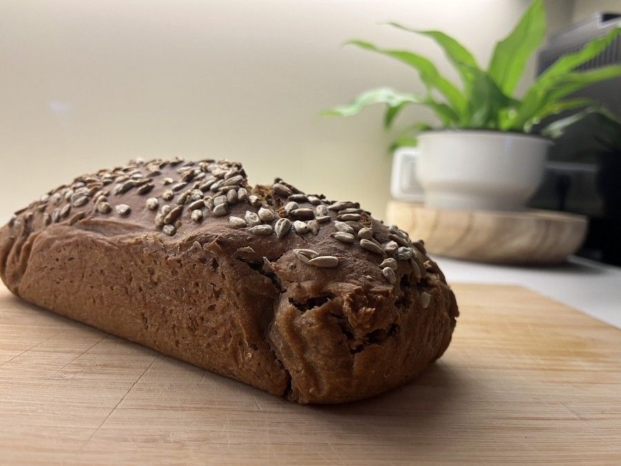 Ein frisch gebackenes Brot, mit Sonnenblumenkernen bestreut, liegt auf einem Holzbrett vor einem modernen Ambiente.