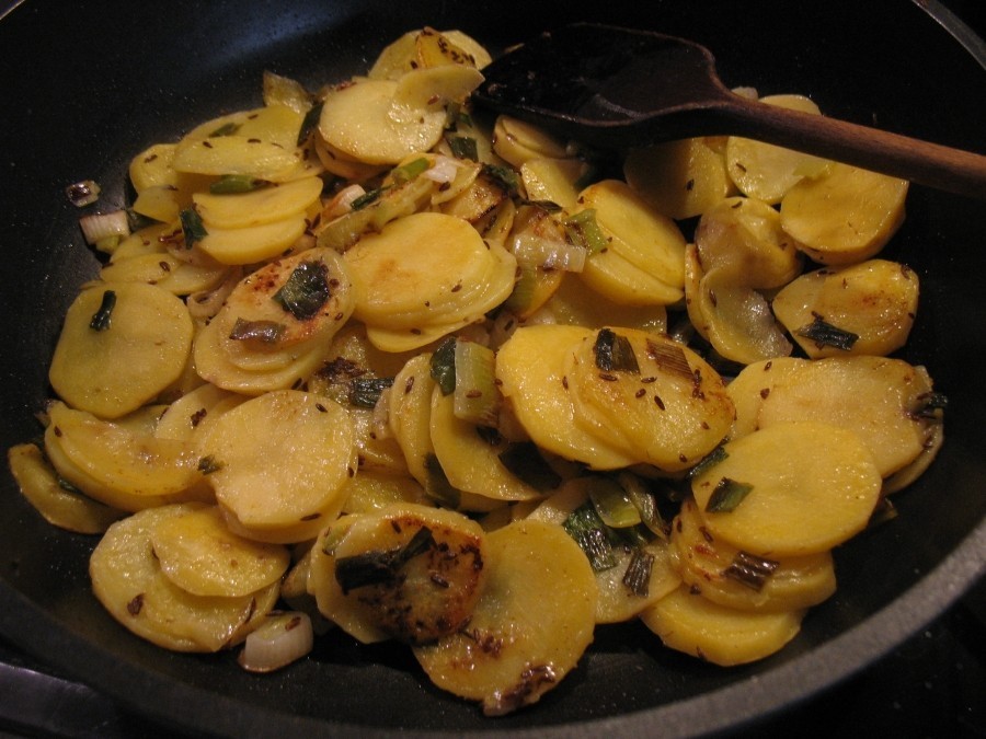 In einer Pfanne werden die Zwiebeln kurz angedünstet, dann die Kartoffelscheiben dazugegeben, mit Salz und Kümmel gewürzt und leicht goldbraun angebraten.  