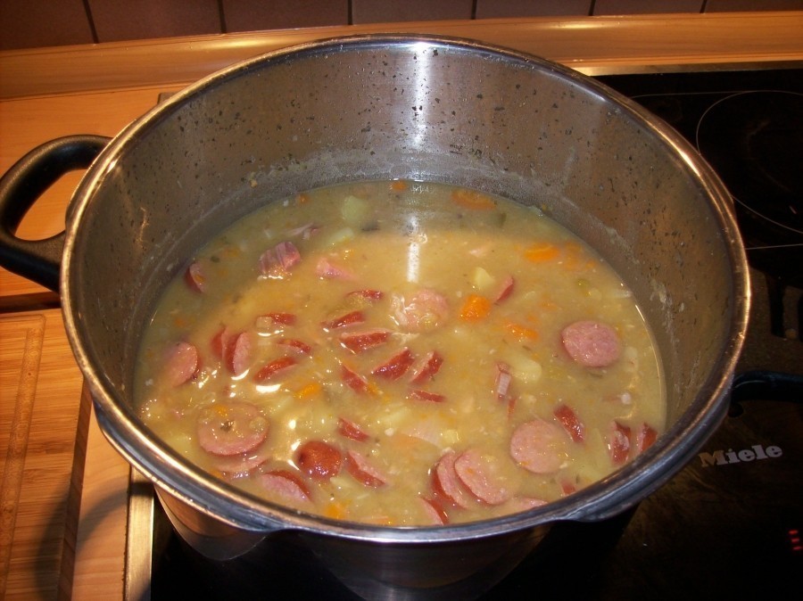 Lässt sich der Kochtopf nun öffnen, wird die Erbsensuppe erst einmal gut umgerührt und sodann kommen die Mettwürstchen-Scheiben und das magere Eisbein-Fleisch wieder in die Suppe.