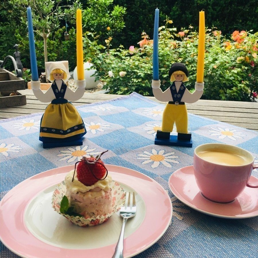 Ein schwedisches Eistörtchen mit Erdbeeren steht auf einem blauen Tisch, umgeben von Kerzen und einer Tasse Kaffee in einem Garten.