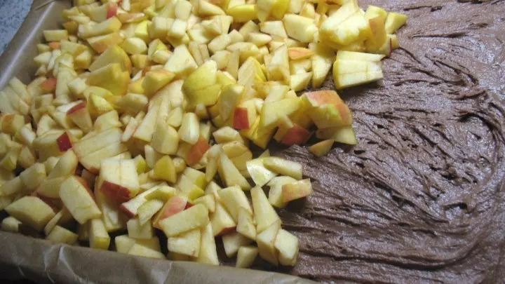 Der Teig wird gleichmäßig auf dem mit Backpapier ausgelegten Blech und darüber die Apfelstückchen und Rosinen verteilt.  