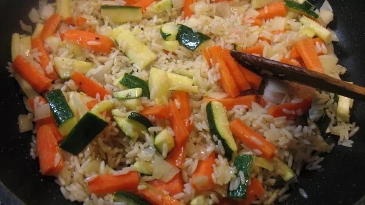 In einer großen Pfanne wird zunächst der gekochte Reis angebraten, dann fügt man die Karotten- und Zucchinistifte und den Knoblauch hinzu und das Ganze wird weiter gebraten.