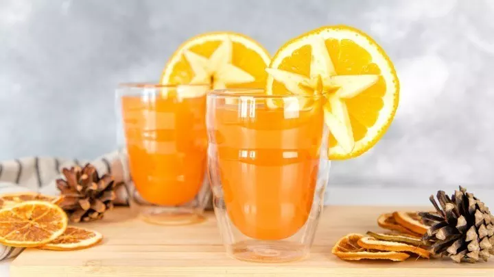 Zwei Gläser mit Hot Aperol und Orangensaft stehen auf einem Holzbrett, dekoriert mit getrockneten Orangenscheiben und Tannenzapfen.