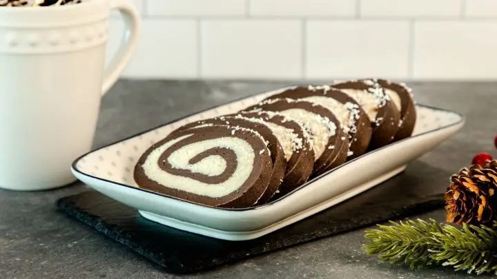 No Bake Cookies aus einer Kokos-Rum-Rolle liegen ordentlich auf einer Platte, umgeben von einer Kaffeetasse und winterlicher Dekoration.