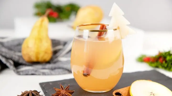 Ein Glas Birne-Zimt-Mocktail steht auf schwarzem Tisch, dekoriert mit Zimtstange und Birnenstück, umgeben von Birnen.