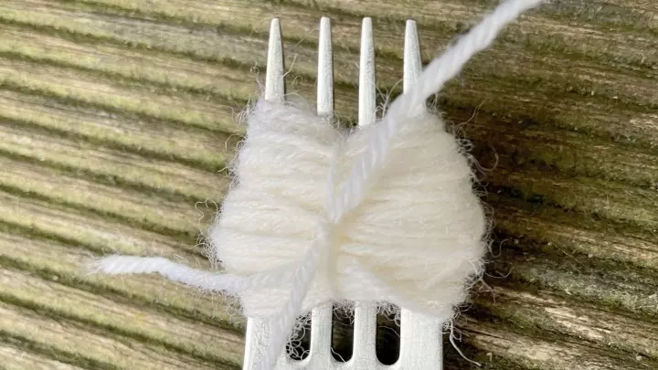 Weiße Wolle um eine Gabel gewickelt. Ein reißfester Faden wurde am mittleren Zinken der Gabel eingezogen und die aufgewickelte Wolle zusammengebunden.