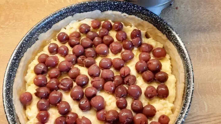 Der noch ungebackene Kuchen wird mit Kirschen belegt. Daneben steht eine Edelstahlschüssel mit Streusel bereit.