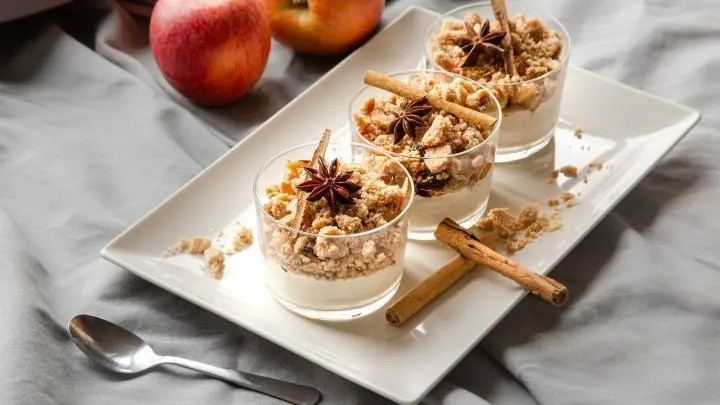 Drei Gl&auml;ser Bratapfel-Crumble auf einem wei&szlig;en Teller mit Zimtstangen, Sternanis dekoriert; zwei &Auml;pfel liegen daneben.