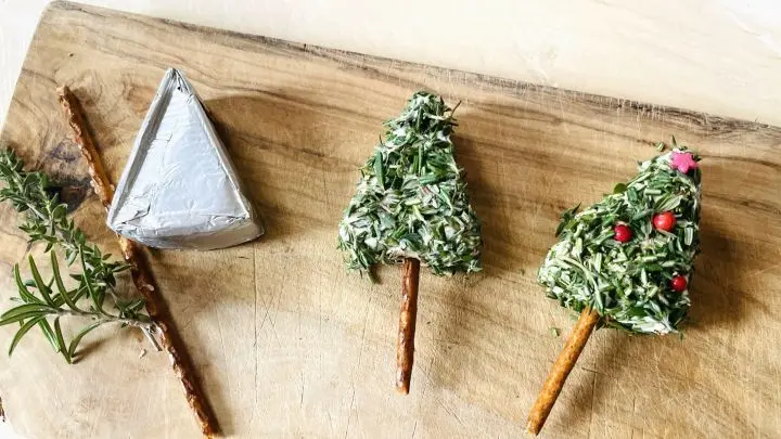 Käse-Tannenbäumchen aus Schmelzkäse und Camembert mit Kräutern bestreut und angerichtet auf einem Brettchen.