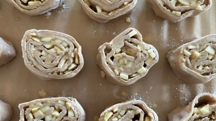 Schnecken aus Teig sind auf einem Backpapier ausgelegt. Sie zeigen Apfelstücke und Zimt, bereit zum Backen.