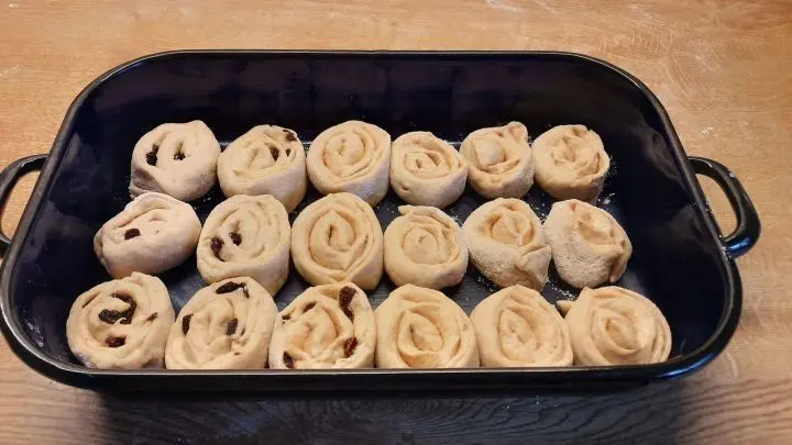 Hefeteigschnecken mit Rosinen und Zimt ruhen in einer rechteckigen schwarzen Backform auf einem Holztisch.